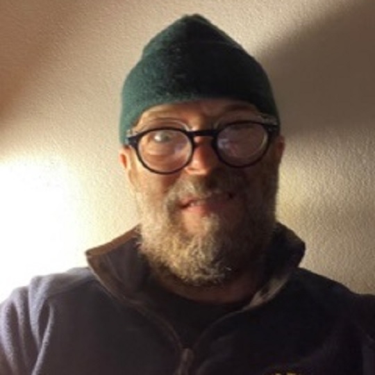 Headshot of Luke Temby wearing a green beanie and glasses and pulling a silly face, he has a grey beard and blue top